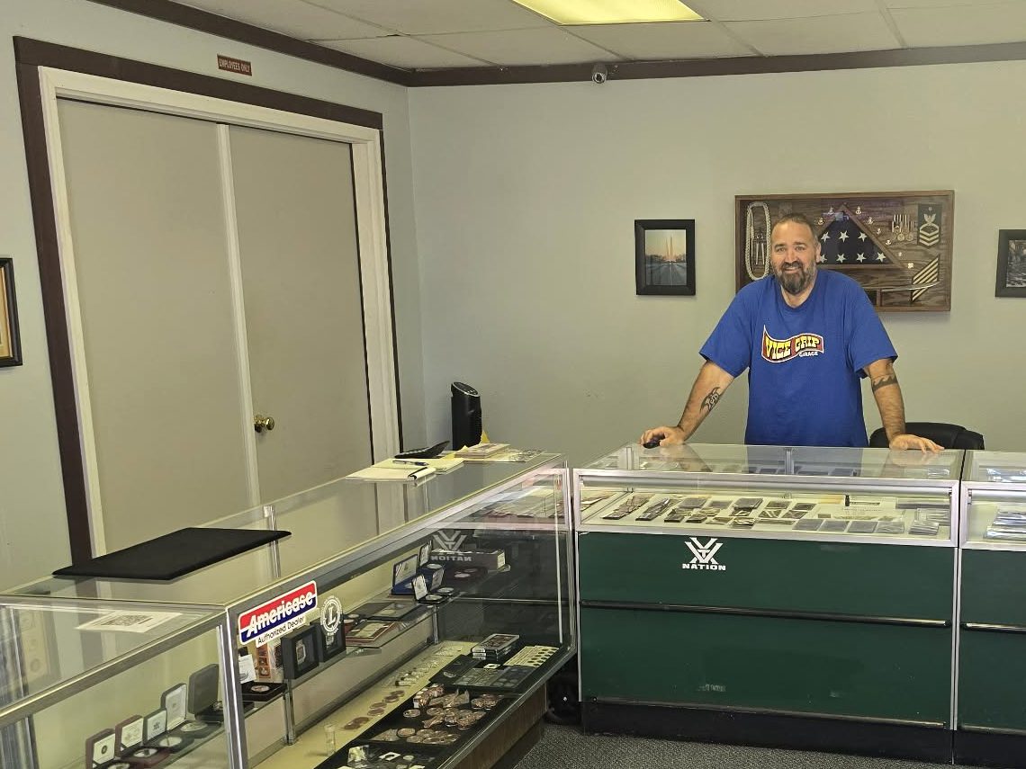 PJ Potter behind the counter at UP North Coins & Bullion, with his Coast Guard shadow box on the wall
